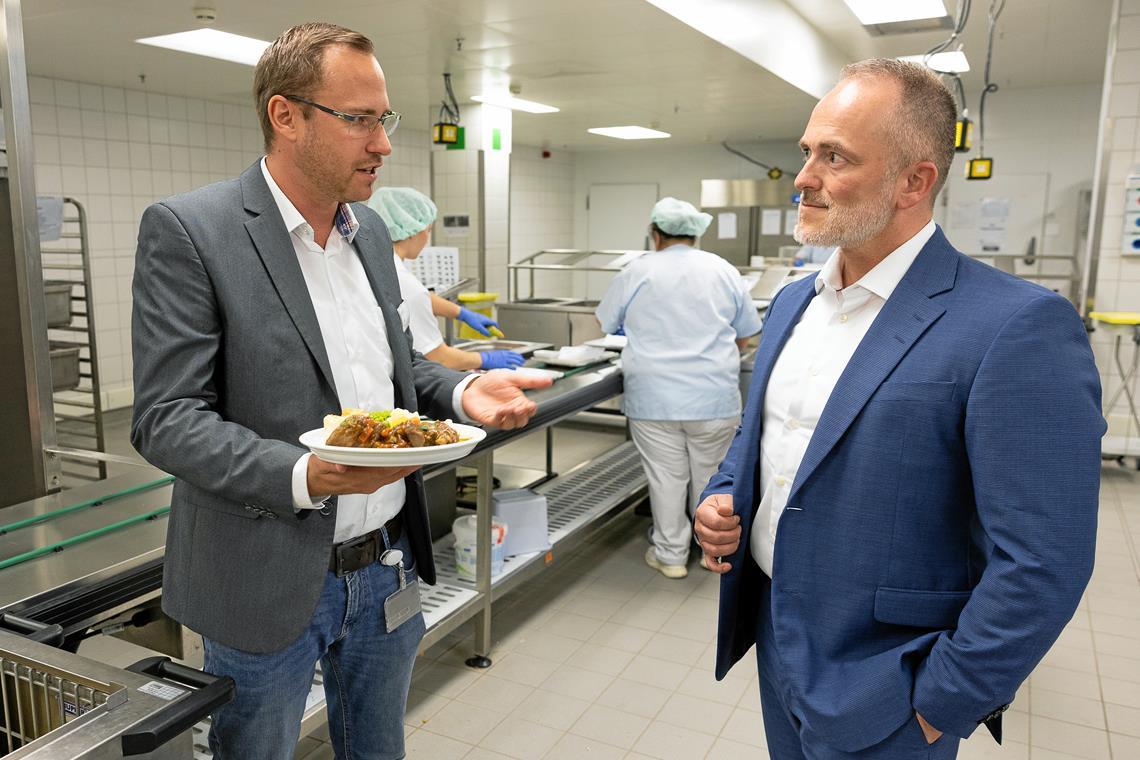 Benjamin Haller, der Leiter des Verpflegungsmanagements, erklärt Kliniken-Geschäftsleiter André Mertel (rechts), dass die Speisen kalt portioniert und erst später erwärmt werden. Fürs Foto stehen sie ausnahmsweise kurz ohne Haube im Küchenbereich. Fotos: Alexander Becher