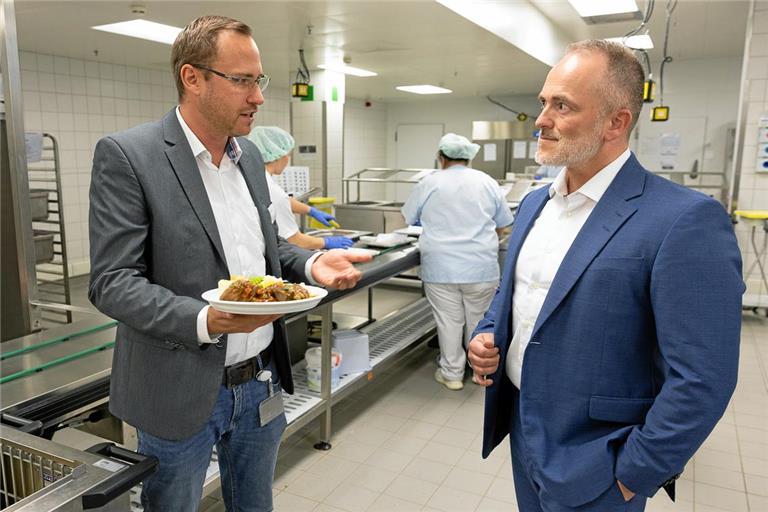 Benjamin Haller, der Leiter des Verpflegungsmanagements, erklärt Kliniken-Geschäftsleiter André Mertel (rechts), dass die Speisen kalt portioniert und erst später erwärmt werden. Fürs Foto stehen sie ausnahmsweise kurz ohne Haube im Küchenbereich. Fotos: Alexander Becher