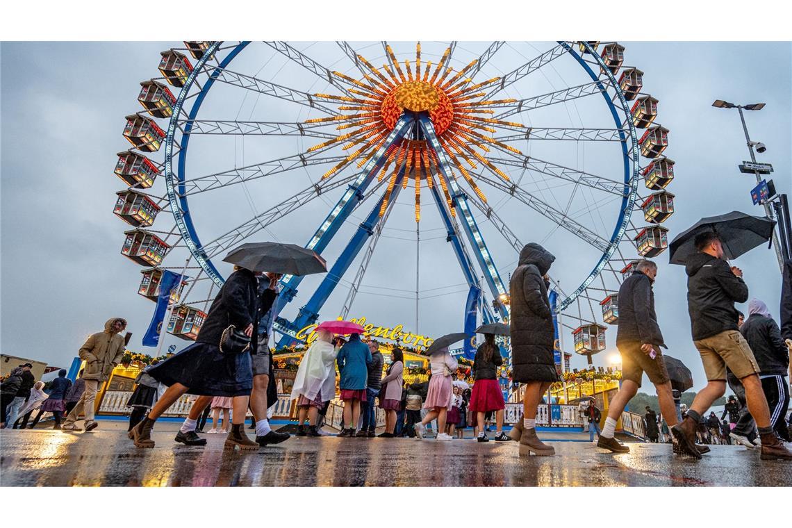Besucher des Oktoberfestes in München gehen im Regen über das Festgelände vor dem Riesenrad.