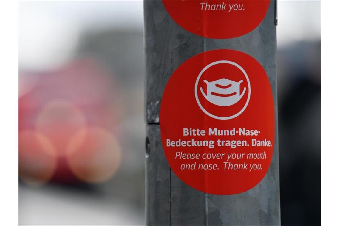 „Bitte Mund-Nase-Bedeckung tragen“ steht in deutscher und englischer Sprache auf einem Aufkleber am Bahnsteig des Westbahnhofs i Jena. Foto: Martin Schutt/dpa-Zentralbild/dpa