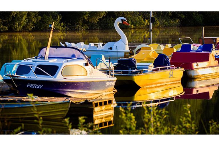 Boote, darunter ein Tretboot in Schwan-Optik, liegen im Licht der aufgehenden Sonne an einem Steg im Altwarmbüchener See.