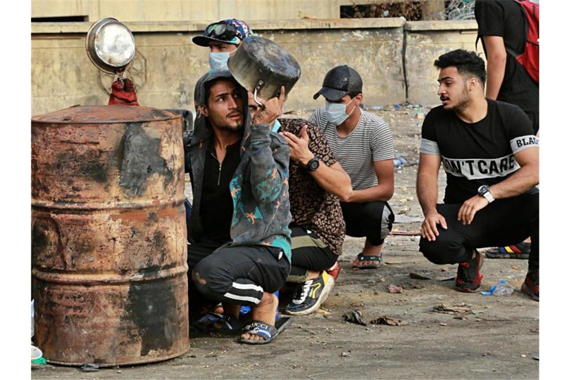 Erneut Proteste im Irak: 63 Menschen sterben Demonstranten hocken in Bagdad hinter Fässern und benutzen Töpfe und Pfannen als Kopfschutz, während Sicherheitskräfte den Zugang zur Grünen Zone blockieren. Foto: Hadi Mizban/AP/dpa