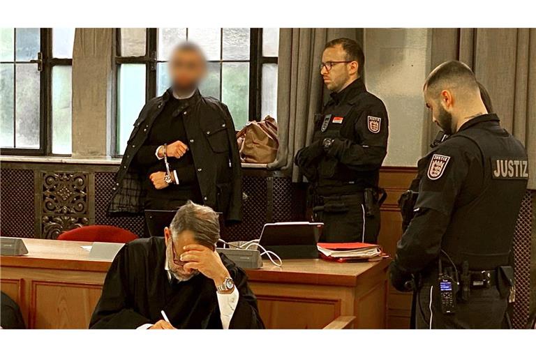 Der Angeklagte (hinten links) steht in Handschellen im Verhandlungssaal im Landgericht neben Justizvollzugsbeamten.