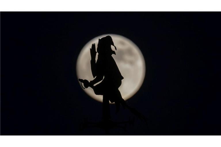 Der fast vollständige Mond steht hinter einer Wetterfahne in Form von einer auf einem Besen reitenden Hexe.