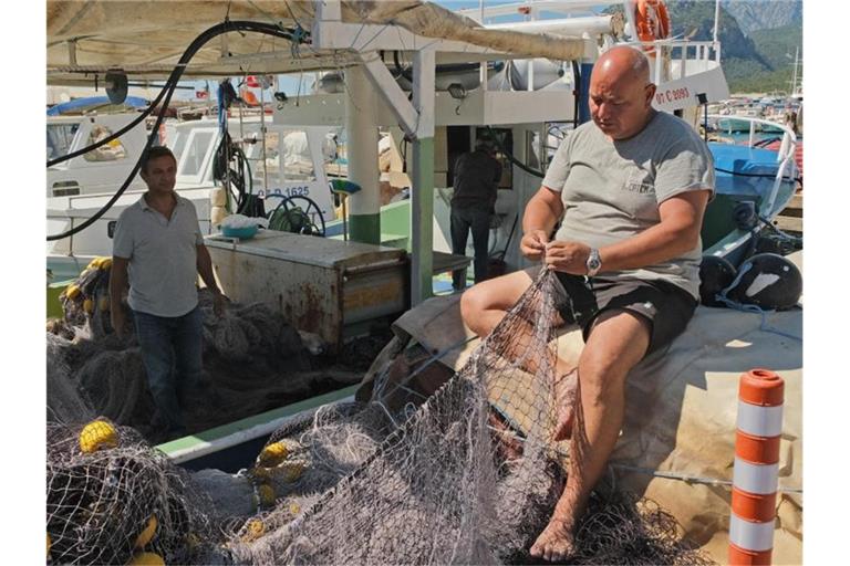 Der Leiter der Fischerei-Kooperative in Antalya, Cengiz Balta, steht auf einem Boot, während ein weiteres Kooperativen-Mitglied im Hafen ein Netz flickt. Foto: Anne Pollmann/dpa