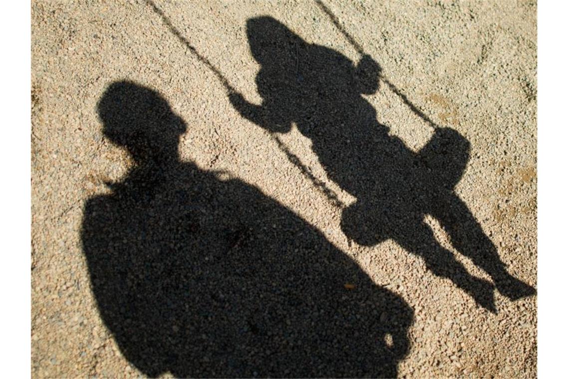 Der Schatten von einem Mann und einem schaukelnden Kind fallen auf Sand auf einem Spielplatz. Foto: Julian Stratenschulte/dpa