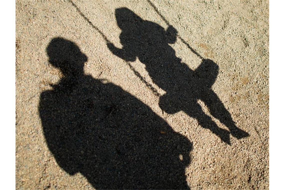 Der Schatten von einem Mann und einem schaukelnden Kind sind im Sand auf einem Spielplatz zu sehen. Foto: Julian Stratenschulte/dpa/Archivbild/dpa/Illustration