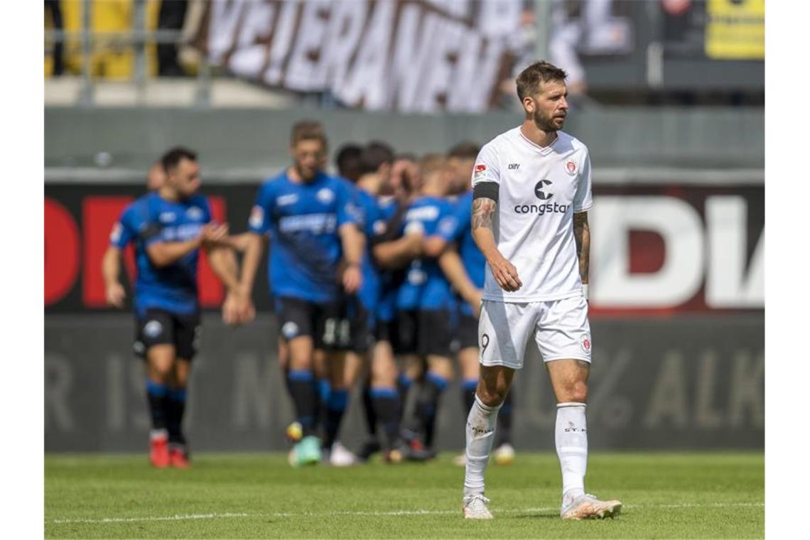 Die Paderborner bejubeln im Hintergrund das Tor zum 2:1, St. Paulis Guido Burgstaller läuft im Vordergrund enttäuscht über den Platz. Foto: David Inderlied/dpa