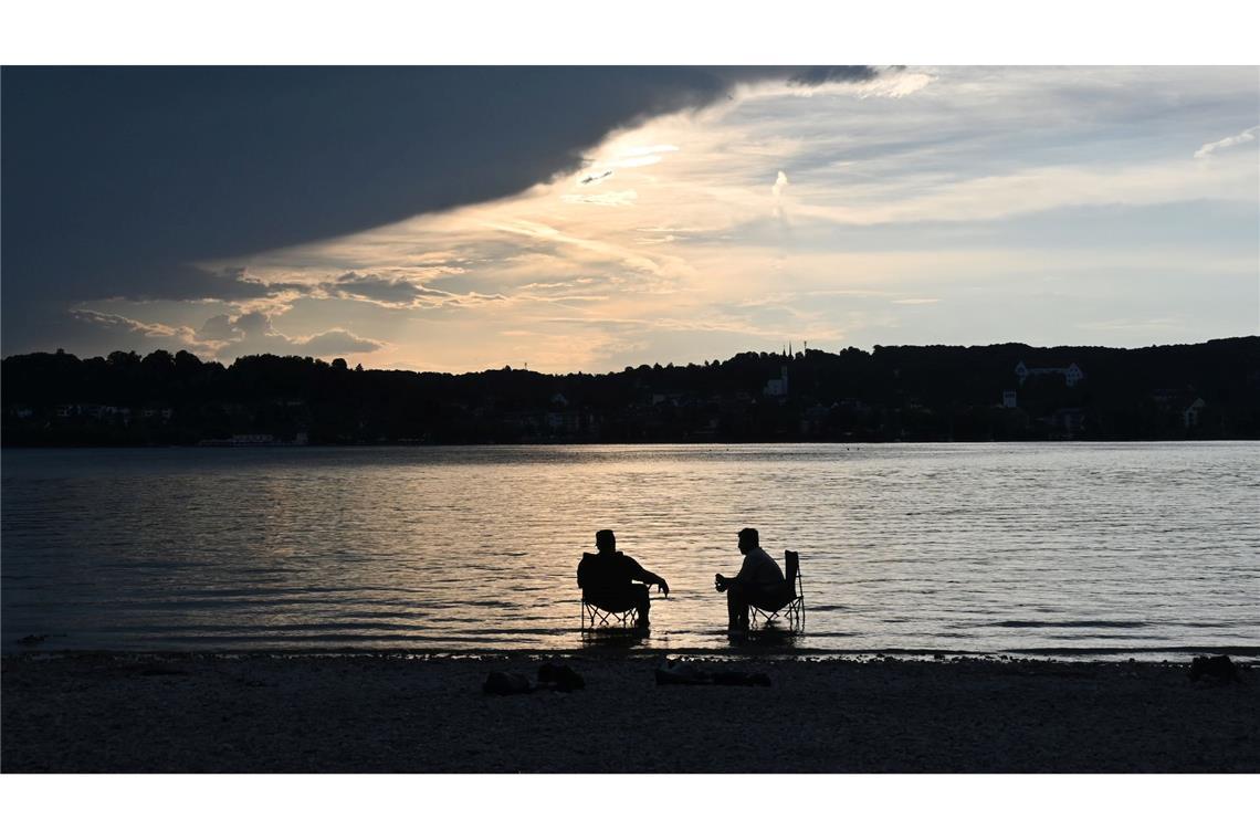 Bilder des Tages Dunkle Wolken ziehen über den Starnberger See während zwei Männer mit Campingstühlen im Wasser sitzen.