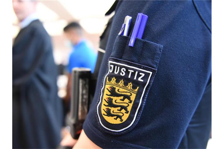 Ein Angeklagter steht im Gerichtssaal in der Messe Ulm, während im Vordergrund eine Justizmitarbeiterin steht, deren Justiz-Logo zu sehen ist. Foto: Felix Kästle/dpa/Archivbild