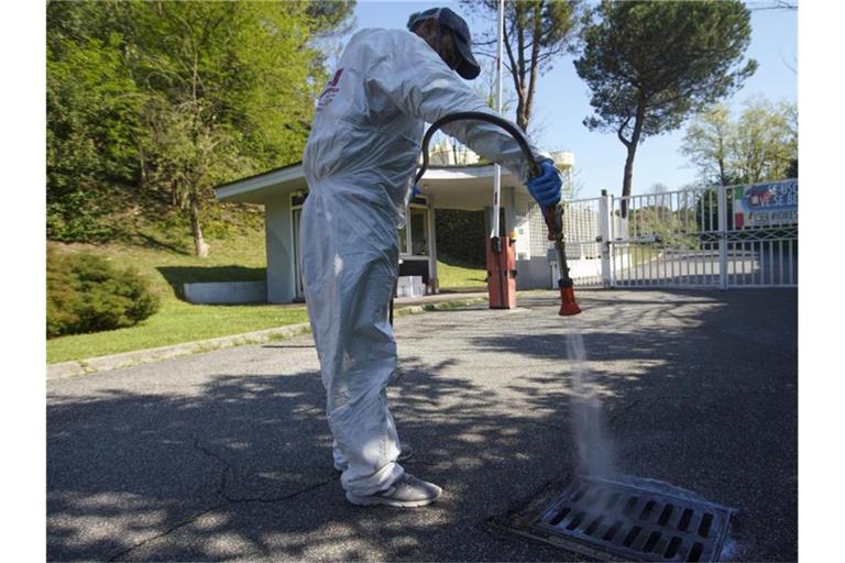 Ein Arbeiter in einem Schutzanzug sprüht Desinfektionsmittel auf ein Kanalisationsgitter in einem Privatviertel Roms. Italien bereitet sich auf eine teilweise Wiedereröffnung nach den langen Ausgangsbeschränkungen vor. Foto: Andrew Medichini/AP/dpa