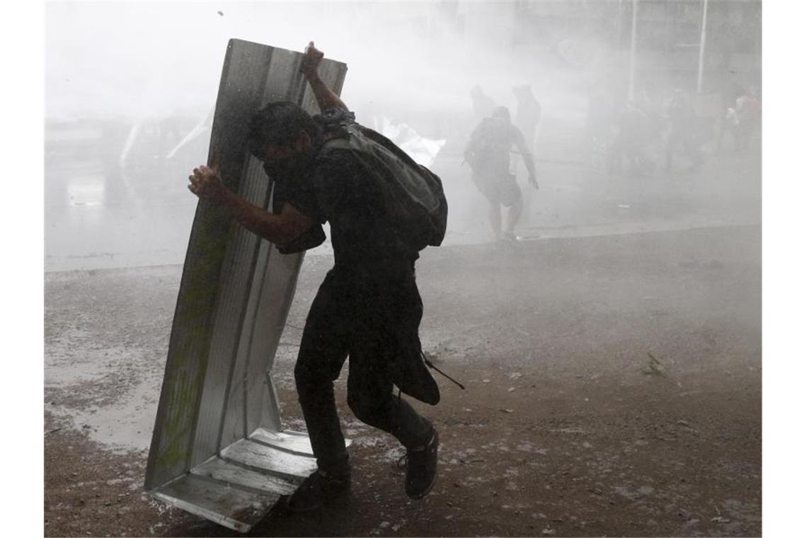 Ein Demonstranten schützt sich mit einem Stück Blech vor einem Wasserwerfer der chilenischen Polizei. Foto: Esteban Felix/AP/dpa