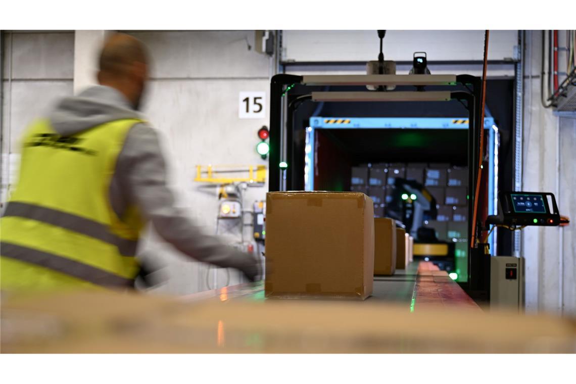 Ein DHL-Mitarbeiter nimmt Pakete in Emfpang - im Hintergrund ist ein Roboter zu sehen, der die Pakete aus einem Container nimmt und sie auf ein Förderband legt. (Archivbild)