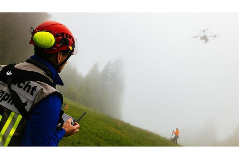 Ein Drohnenpilot der Bergwacht sieht zu, wie ein Flughelfer der Bergwacht eine Übungslast von der Seilwinde einer Lastendrohne abhängt.