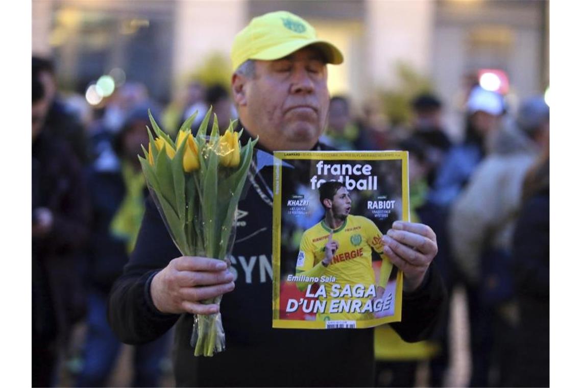 Ein Fan des FC Nantes hält gelbe Blumen und ein Fußballmagazin mit dem verschollenen Fußballspieler Emiliano Sala auf dem Cover. Foto: David Vincent/AP