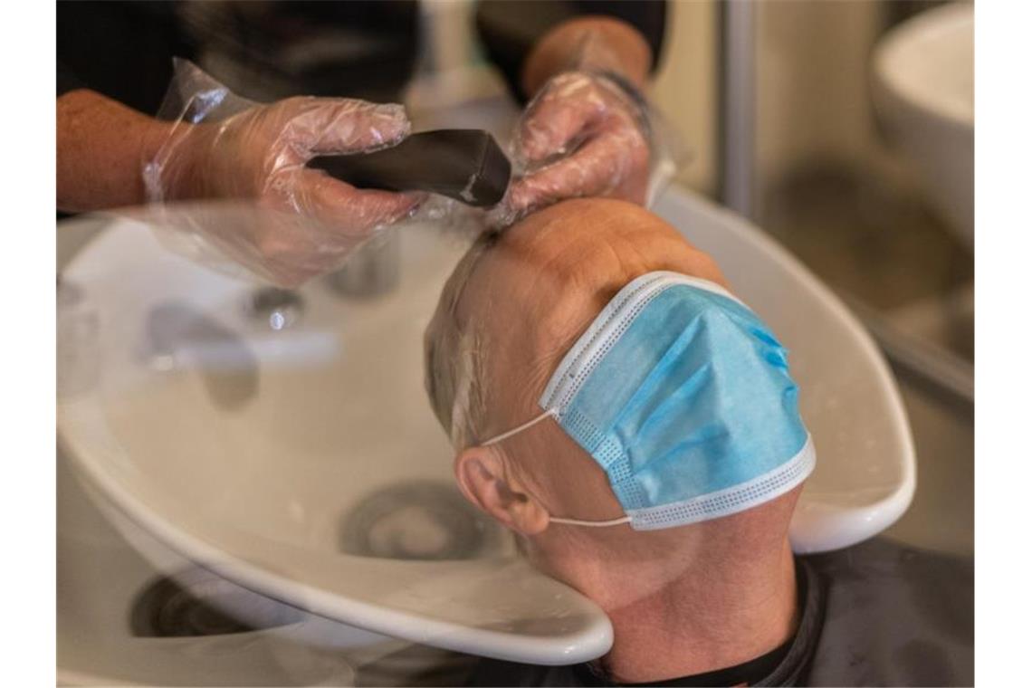 Ein Friseurmeister wäscht einem Kunden mit Mund- und Nasenschutz vor dem Schnitt die Haare. Foto: Robert Michael/dpa-Zentralbild/dpa