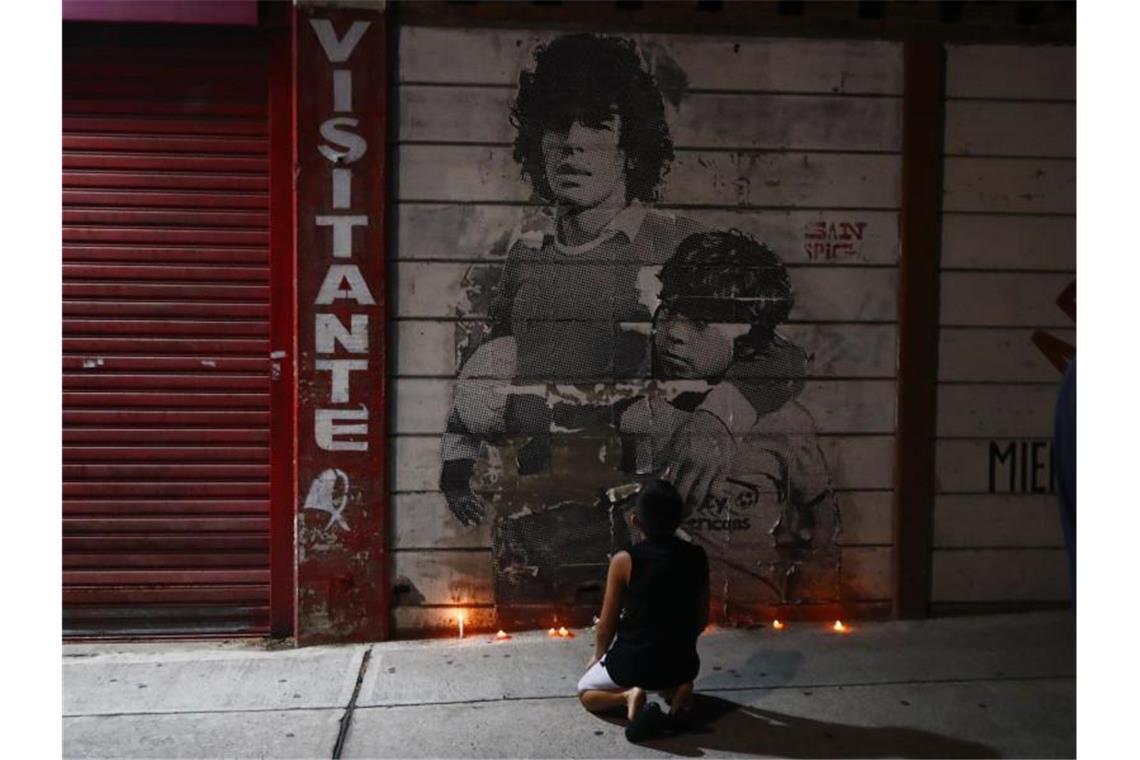 Ein Junge kniet vor einem mit Kerzen dekorierten Wandbild von Diego Maradona in Argentinien. Foto: Marcos Brindicci/AP/dpa