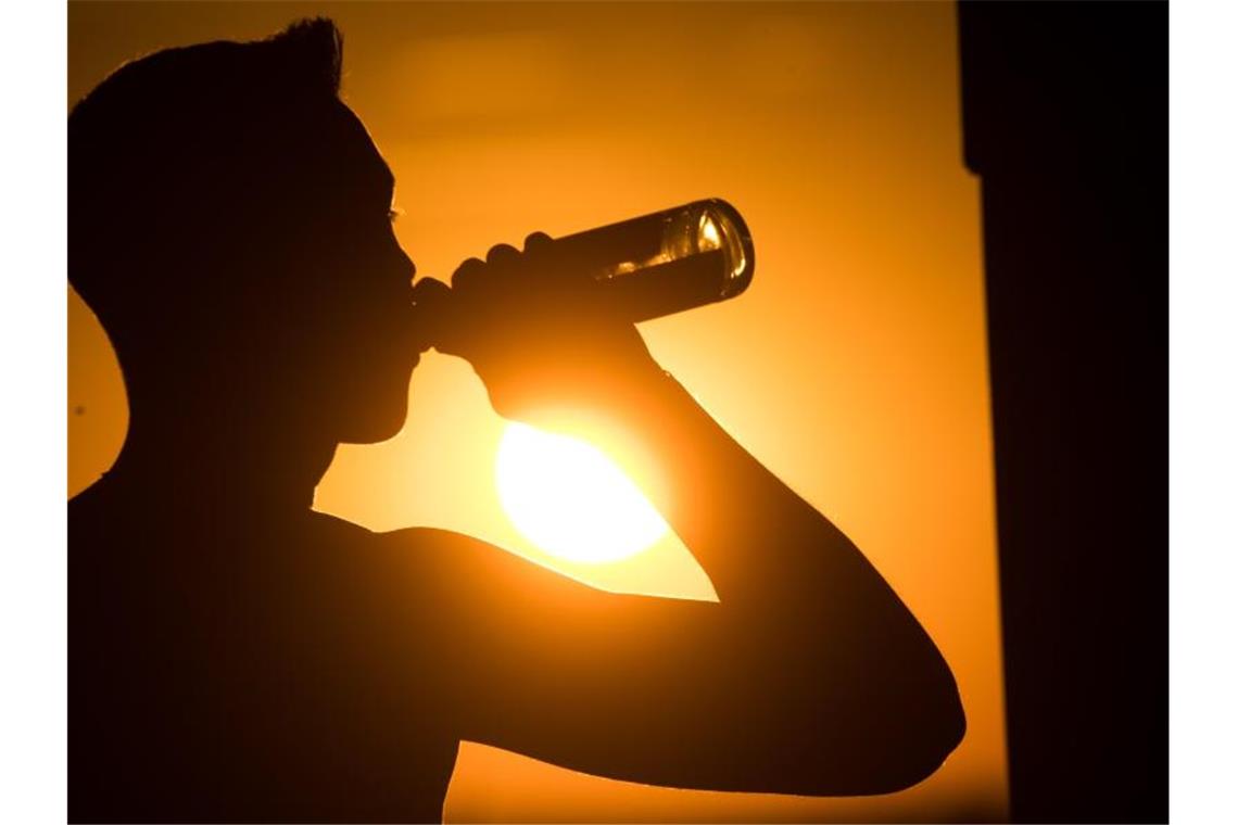 Ein junger Mann sitzt auf dem Kronsberg, und trinkt Bier, während am Horizont die Sonne untergeht. Foto: Julian Stratenschulte/dpa