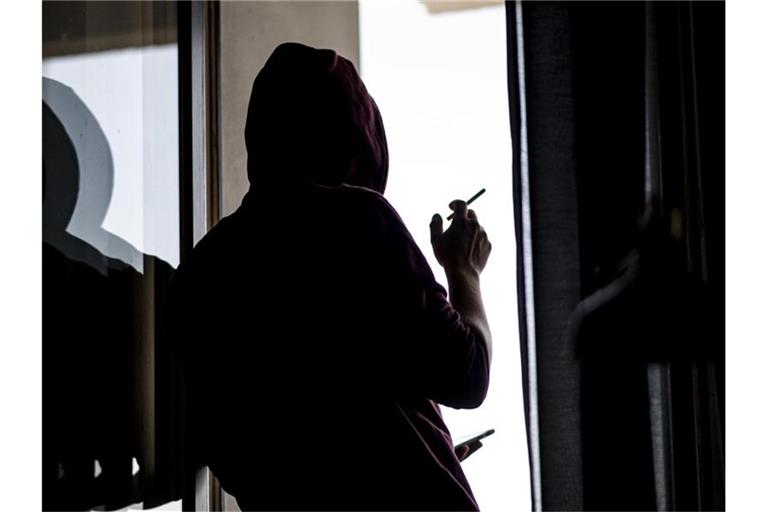 Ein junger Mann steht mit Zigarette und Smartphone an einem Fenster. Foto: Fabian Sommer/dpa/Symbolbild
