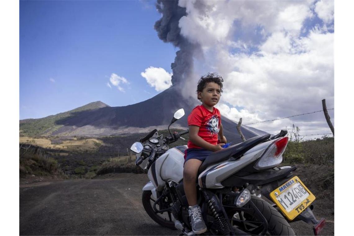 Ein Kind sitzt auf einem Motorrad, während im Hintergrund der Vulkan Pacaya ausbricht. Foto: Santiago Billy/AP/dpa