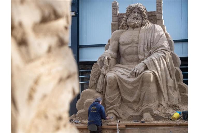 Ein Mann arbeitet an einer Sandskulptur des Göttervaters Zeus auf seinem Thron. Foto: Stefan Sauer/dpa