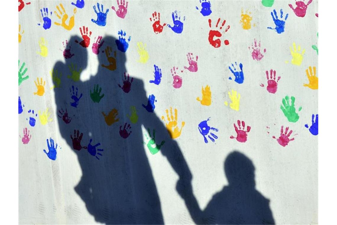 Ein Mann mit einem Kind auf dem Arm und einem an der Hand wirft einen Schatten auf eine mit bunten Handabdrücken bemalte Wand. Foto: Peter Kneffel/dpa/Symbolbild