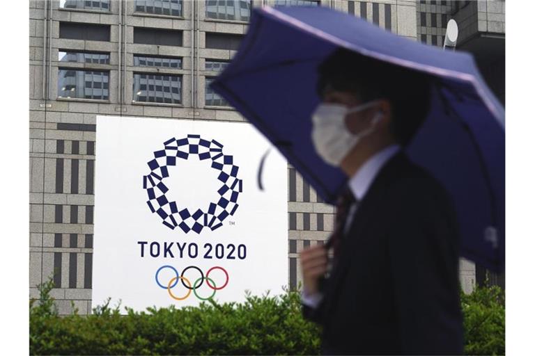 Ein Mann mit Mund-Nasen-Schutz geht im Regen an einem Banner der Olympischen Spiele Tokio 2020 vorbei. Foto: Eugene Hoshiko/AP/dpa