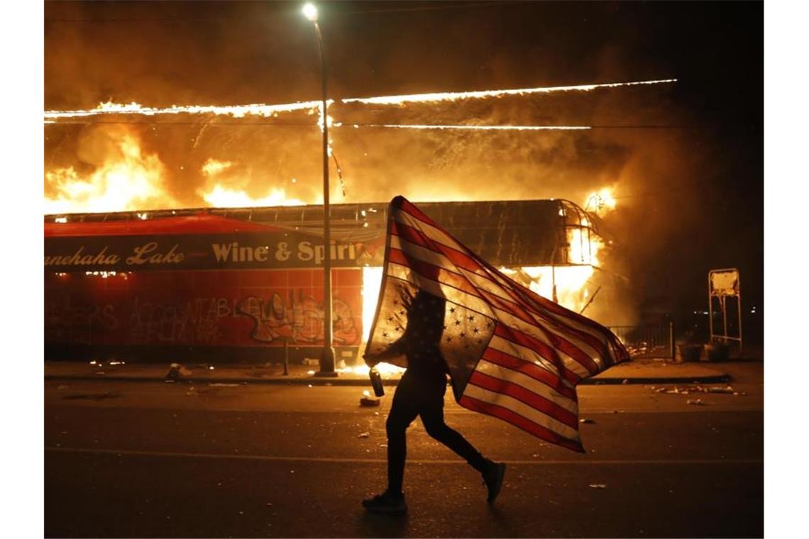 Ein Mann mit umgedrehter US-Flagge vor einem brennenden Gebäude in Minneapolis. Foto: Julio Cortez/AP/dpa