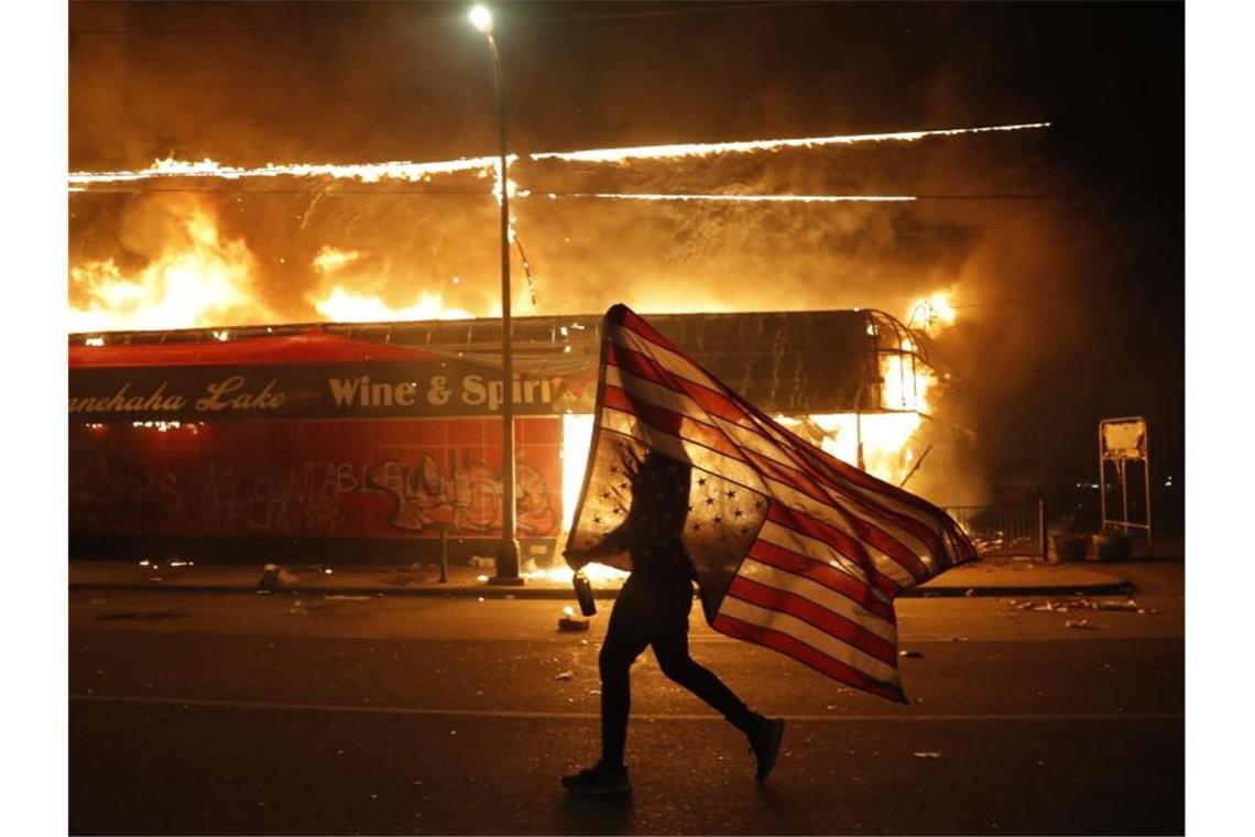 Welle von Protesten und Gewalt erschüttert die USA Ein Mann mit umgedrehter US-Flagge vor einem brennenden Gebäude in Minneapolis. Foto: Julio Cortez/AP/dpa