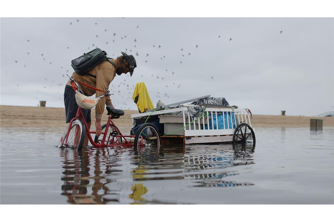 Ein Mann passt seine Ladung auf dem Fahrradanhänger an, um den durch heftigen Regenfällen überfluteten Weg entlang zufahren.