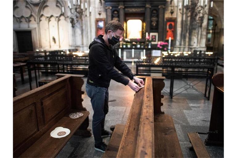 Ein Mitarbeiter der Dombauhütte klebt Aufkleber auf Kirchenbänke im Kölner Dom. Die runden Sticker sollen den Platz markieren, auf den Gottesdienstbesucher sich setzen dürfen. Foto: Federico Gambarini/dpa