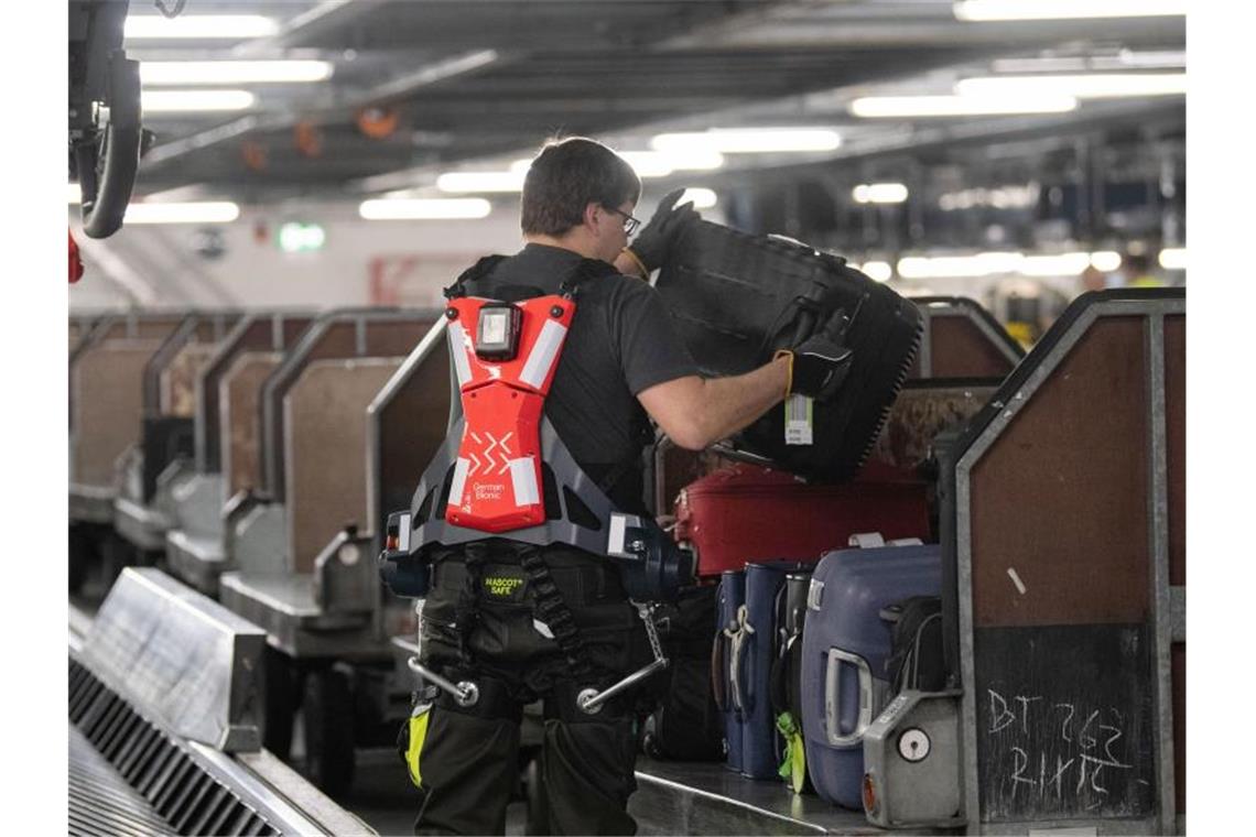 Ein Mitarbeiter des Stuttgarter Flughafens hebt mit Unterstützung eines Exoskeletts einen Koffer auf einen Gepäckwagen. Foto: Marijan Murat/dpa