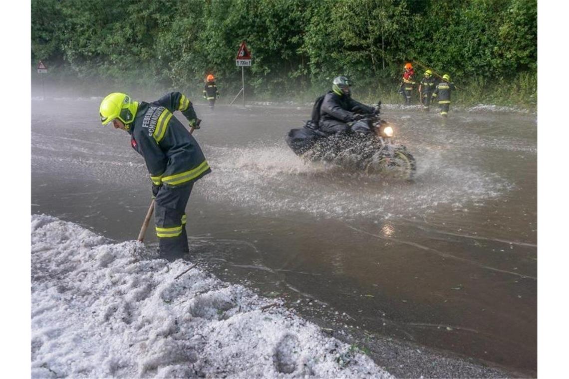 Ein Motorradfahrer fährt nach einem schweren Unwetter über eine überflutete Straße, während Einsatzkräfte der Feuerwehr an den Straßenseiten Hagelkörner wegschippen. Foto: Einsatzdoku/APA/dpa
