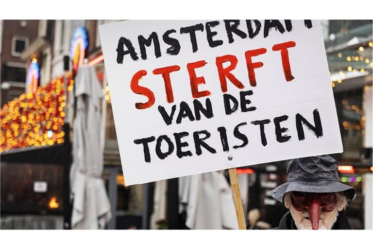 Ein niederländischer Aktivist mit Narrenmaske hält ein  Schild hoch mit der übersetzten Aufschrift: „Amsterdam stirbt an den Touristen“