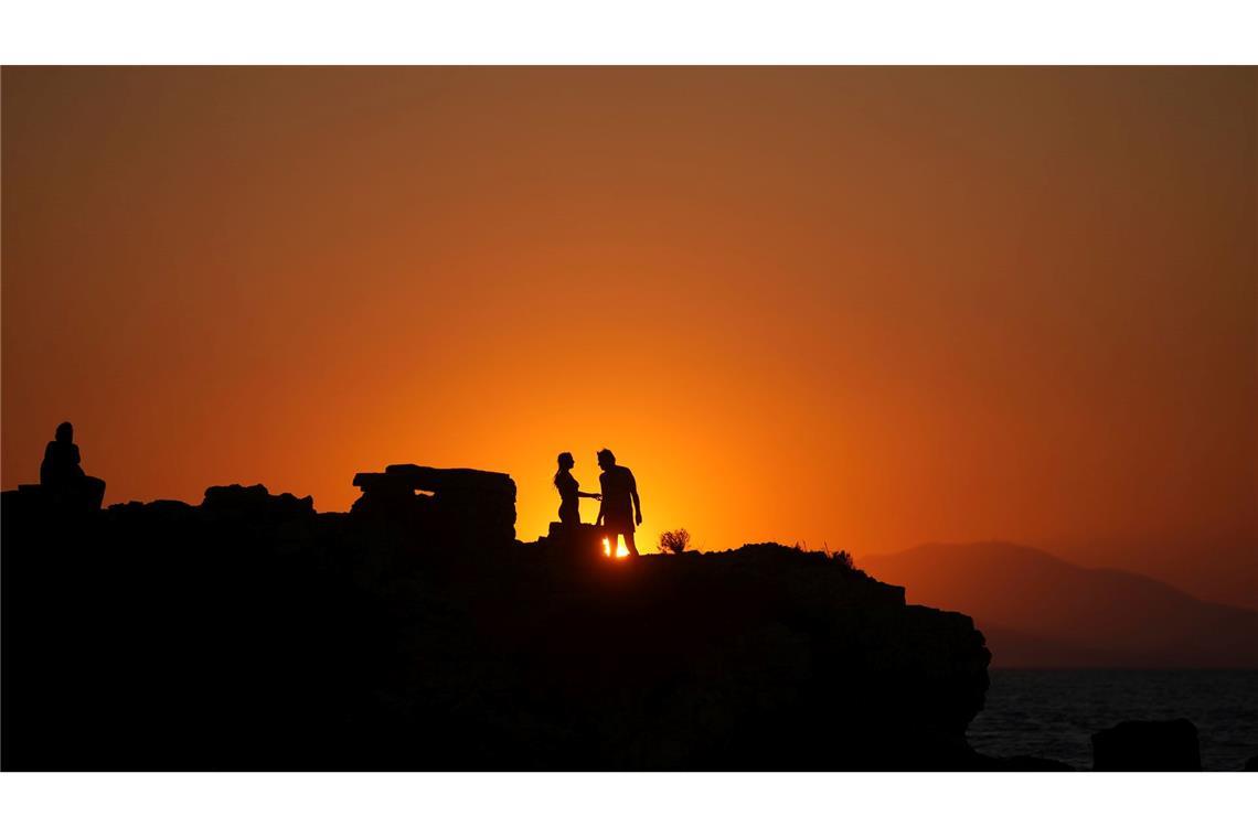 Ein Paar steht auf einem Meeresfelsen, während die Sonne an einem warmen und windigen Tag am Strand von Kavouri im Süden Athens untergeht,