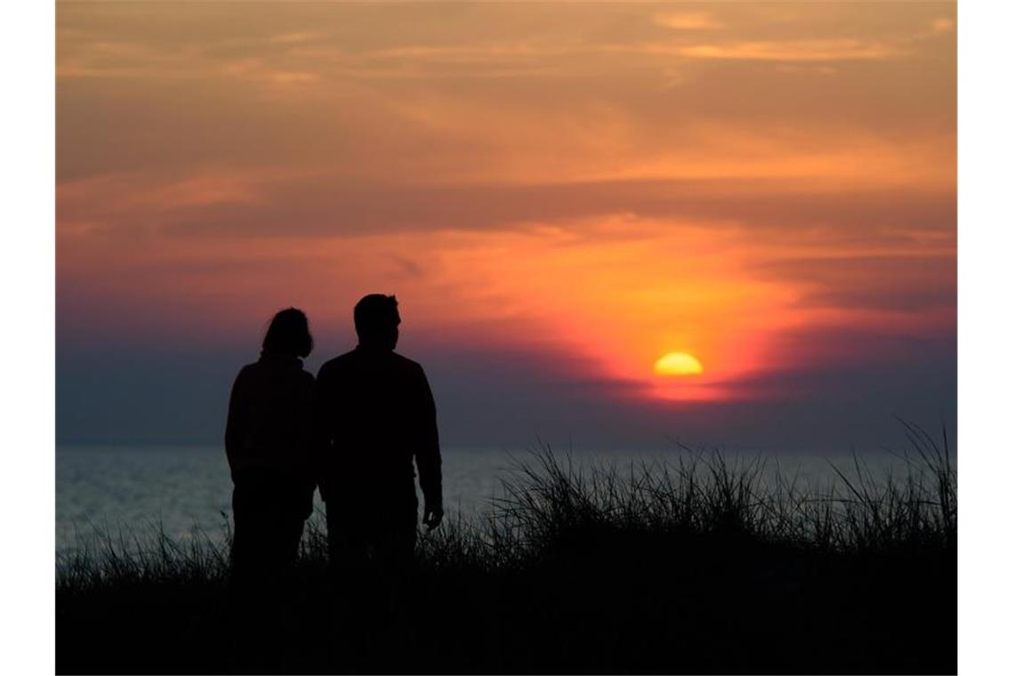 Ein Paar steht während des rötlich-orangenen Sonnenunterganges über der Ostsee an einer Düne am Strand. Foto: Gregor Fischer/dpa