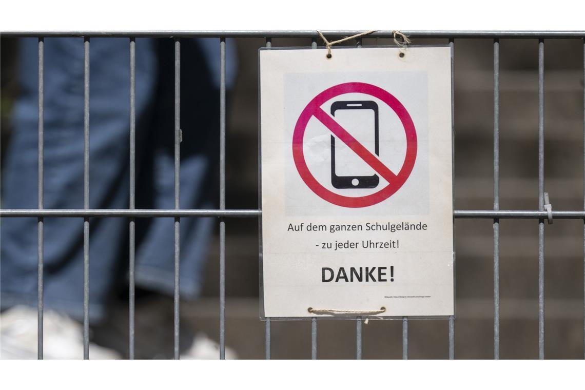 Ein Schild mit einem durchgestrichenen Smartphone und der Aufschrift „Auf dem ganzen Schulgelände - zu jeder Uhrzeit! DANKE!“ hängt am Zugangstor zum Schulhof eines Gymnasiums. (Symbolbild)