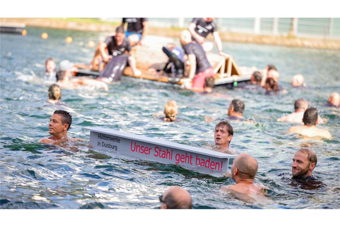 "Unser Stahl geht baden": Stahlarbeiter springen ins Wasser Ein symbolischer Stahlträger mit dem Slogan der Demo schwimmt im Duisburger Innenhafen zwischen zahlreichen Stahlbeschäftigten.