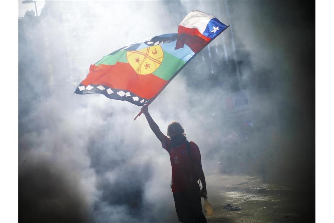 Gipfelabsagen in Chile: Weltklimakonferenz zieht wohl um Ein vermummter Demonstrant weht die Flagge von Chile und der indigenen Gemeinschaft Mapuche mitten im Rauch während eines Protestes für Sozialreformen. Foto: Agenciauno/Agencia Uno/dpa