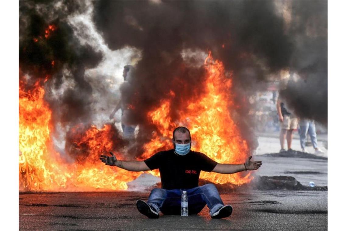 Ein Demonstrant mit Mundschutz sitzt mit ausgestreckten Armen vor einer brennenden Straßenbarrikade. Foto: Marwan Naamani/dpa