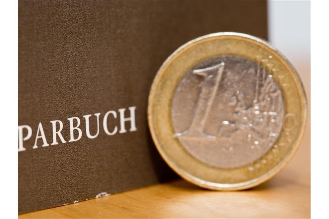 Eine Ein-Euro-Münze steht neben einem Sparbuch. Foto: Daniel Karmann/dpa/Illustration