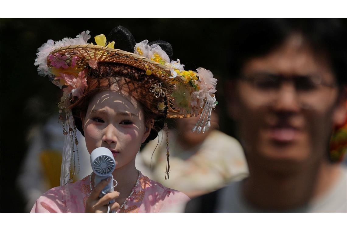 Eine Frau, die einen Hut mit Blumen und ein traditionelles Kostüm trägt, kühlt sich mit einem tragbaren elektrischen Ventilator ab, während Menschen an einem heißen Tag die Verbotene Stadt in Peking besuchen.