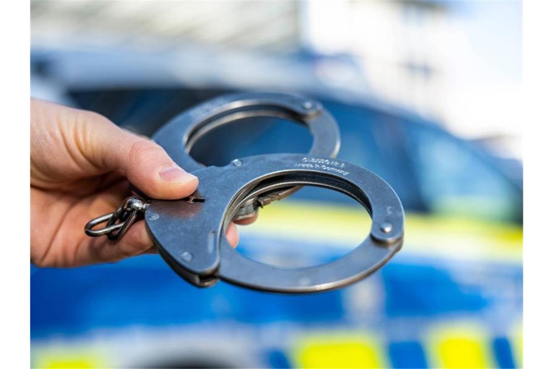 18-Jähriger mit Messer verletzt: Verdächtiger in U-Haft Eine Hand hält Handschellen vor einen Streifenwagen der Polizei. Foto: David Inderlied/dpa/Illustration