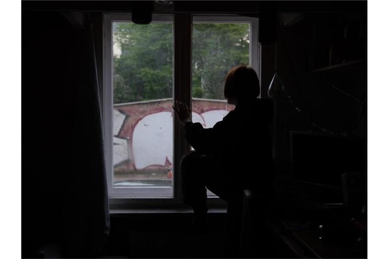 Eine Jugendliche sitzt in einem abgedunkelten Zimmer am Fenster. Foto: Paul Zinken/dpa