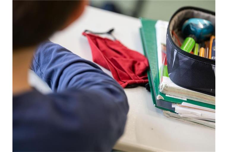 Eine Mund-Nasen-Bedeckung liegt während einer Unterrichtsstunde neben einem Mäppchen und Schulbüchern auf einem Schultisch. Foto: Marijan Murat/dpa/Archivbild