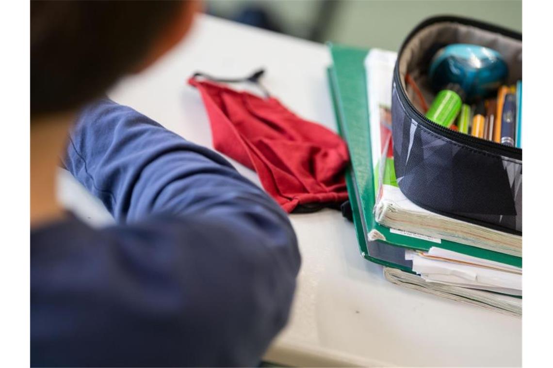 Eine Mund-Nasen-Bedeckung liegt während einer Unterrichtsstunde neben einem Mäppchen und Schulbüchern auf einem Schultisch. Foto: Marijan Murat/dpa/Symbolbild/Archiv