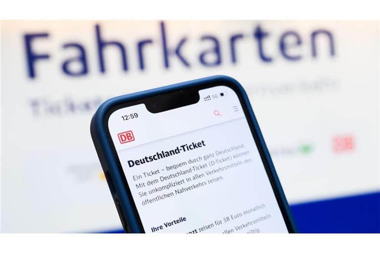 Eine Person hält ein Smartphone in der Hand, auf dessen Display eine Information zum Kauf des Deutschlandtickets zu sehen ist, während sie vor einem Fahrkartenautomaten steht. 