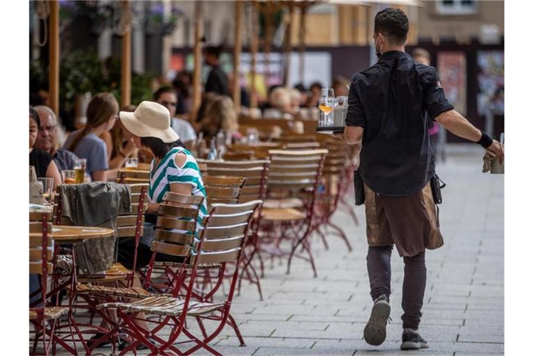 Eine Servicekraft läuft im Außenbereich eines Restaurants an Gästen vorbei. Foto: Christoph Schmidt/dpa