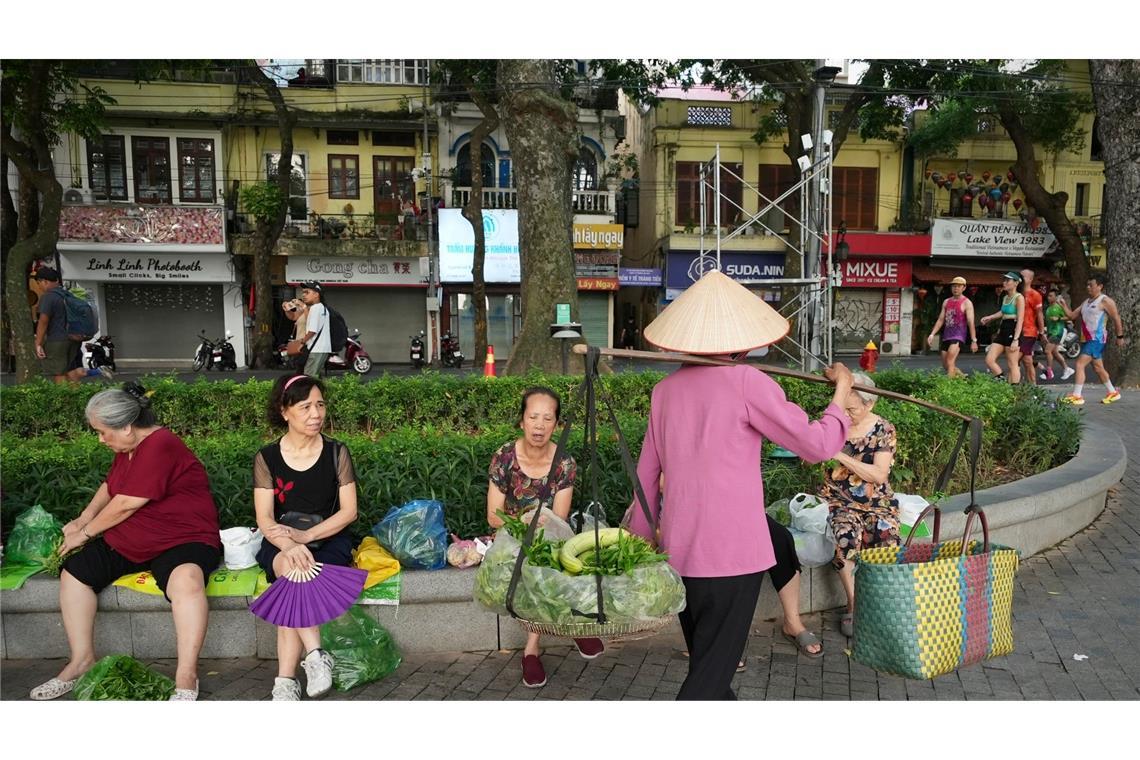 Eine Verkäuferin verkauft Gemüse an Menschen in Hanoi, die in einem Park Morgensport treiben.