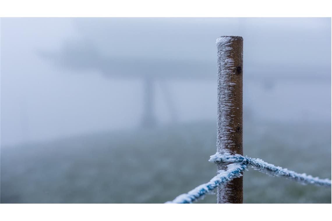 Schnee fällt nachts auf dem Feldberg - am Morgen nur Sturm Eiskristalle hängen an einem Holzpfahl während im Hintergrund die Bergstation der Feldbergbahn zu sehen ist.